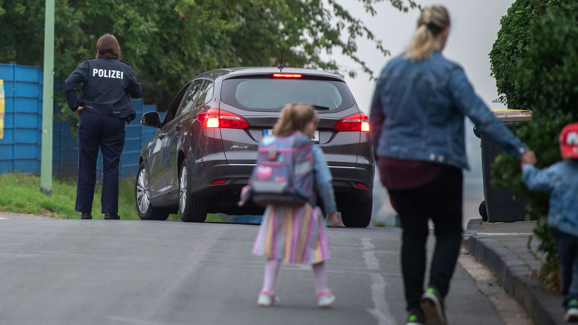 Eine Frau geht mit zwei Kindern über die Straße, im Hintergrund sind ein Auto und eine Polizistin zu sehen.