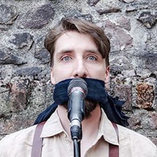 Juri von Stavenhagen hat ein Tuch über den Mund gebunden und steht hinter einen Mikrofon. .