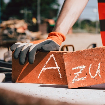 Ein Mann hält in jeder Hand einen Backstein, auf einem steht ein „A“, auf dem anderen der Schriftzug „zubi“.