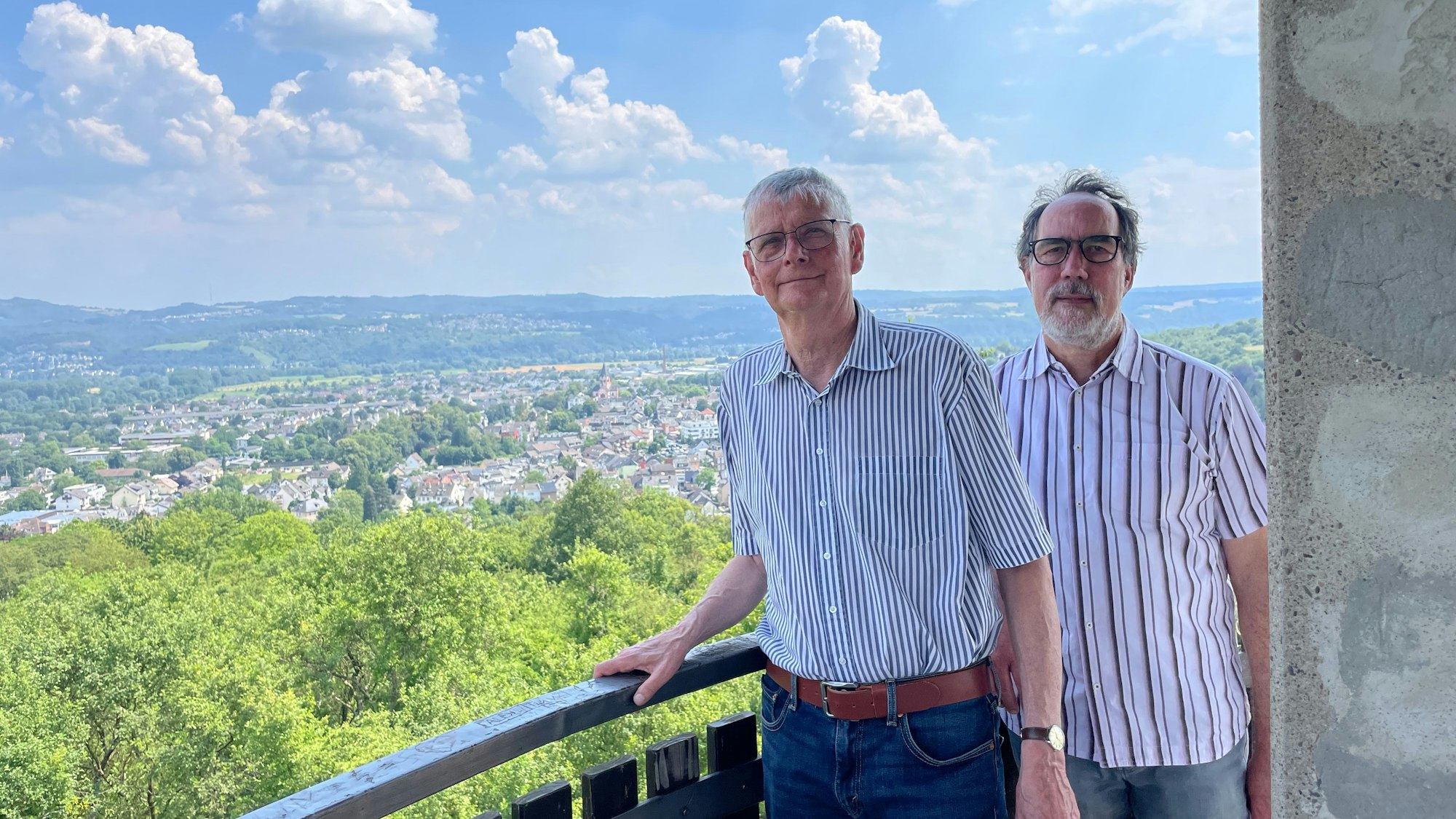 Die beiden Männer stehen an einem Geländer, dahinter ist weithin Landschaft mit Bebauung und Wäldern zu sehen.