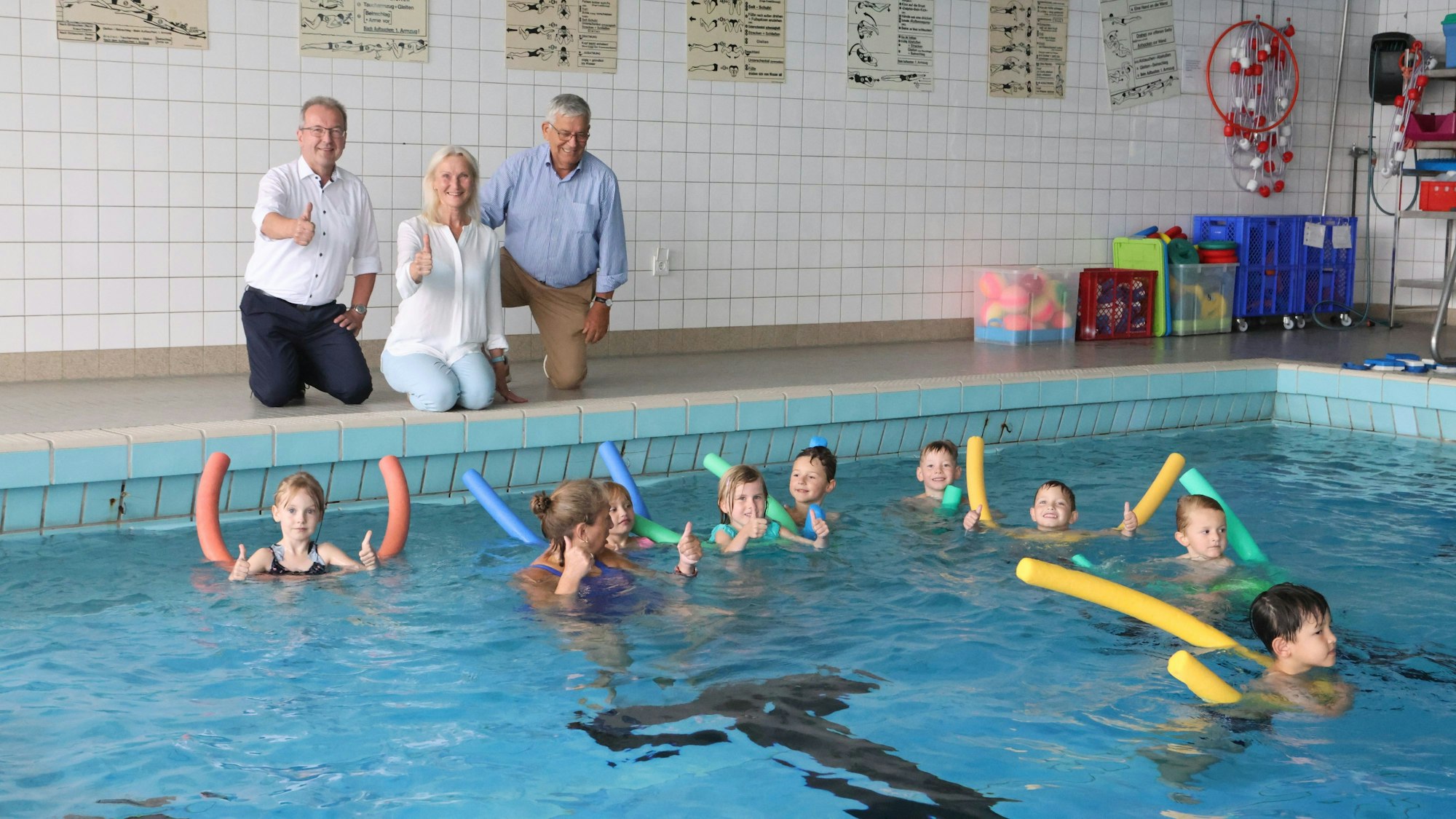 Drei Erwachsene hocken mit erhobenen Daumen neben einem Schwimmbecken, in dem gerade eine Gruppe Kinder das Schwimmen lernt.