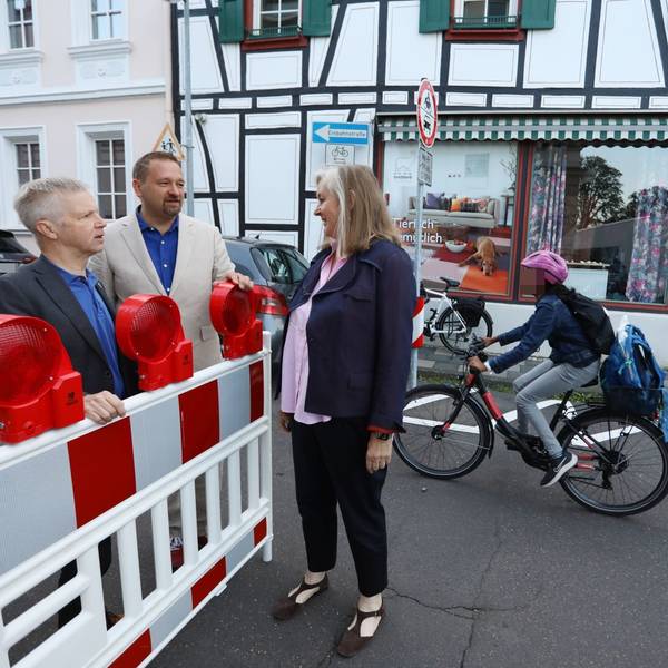 Zwei Männer und eine Frau stehen an einer rot-weißen Sperrbake, im Hintergrund fährt ein Schüler auf dem Fahrrad vorbei.
