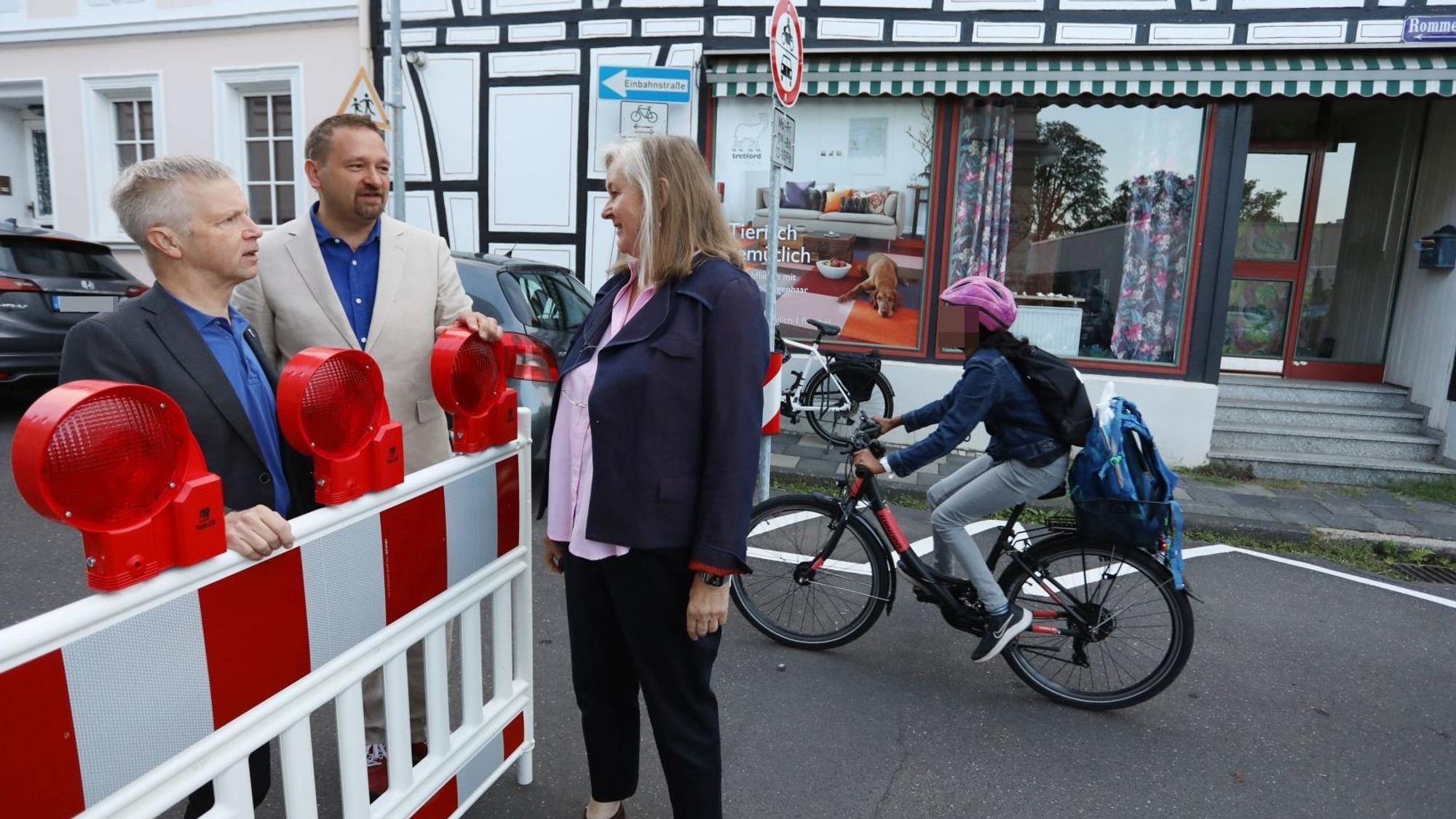 Zwei Männer und eine Frau stehen an einer rot-weißen Sperrbake, im Hintergrund fährt ein Schüler auf dem Fahrrad vorbei.