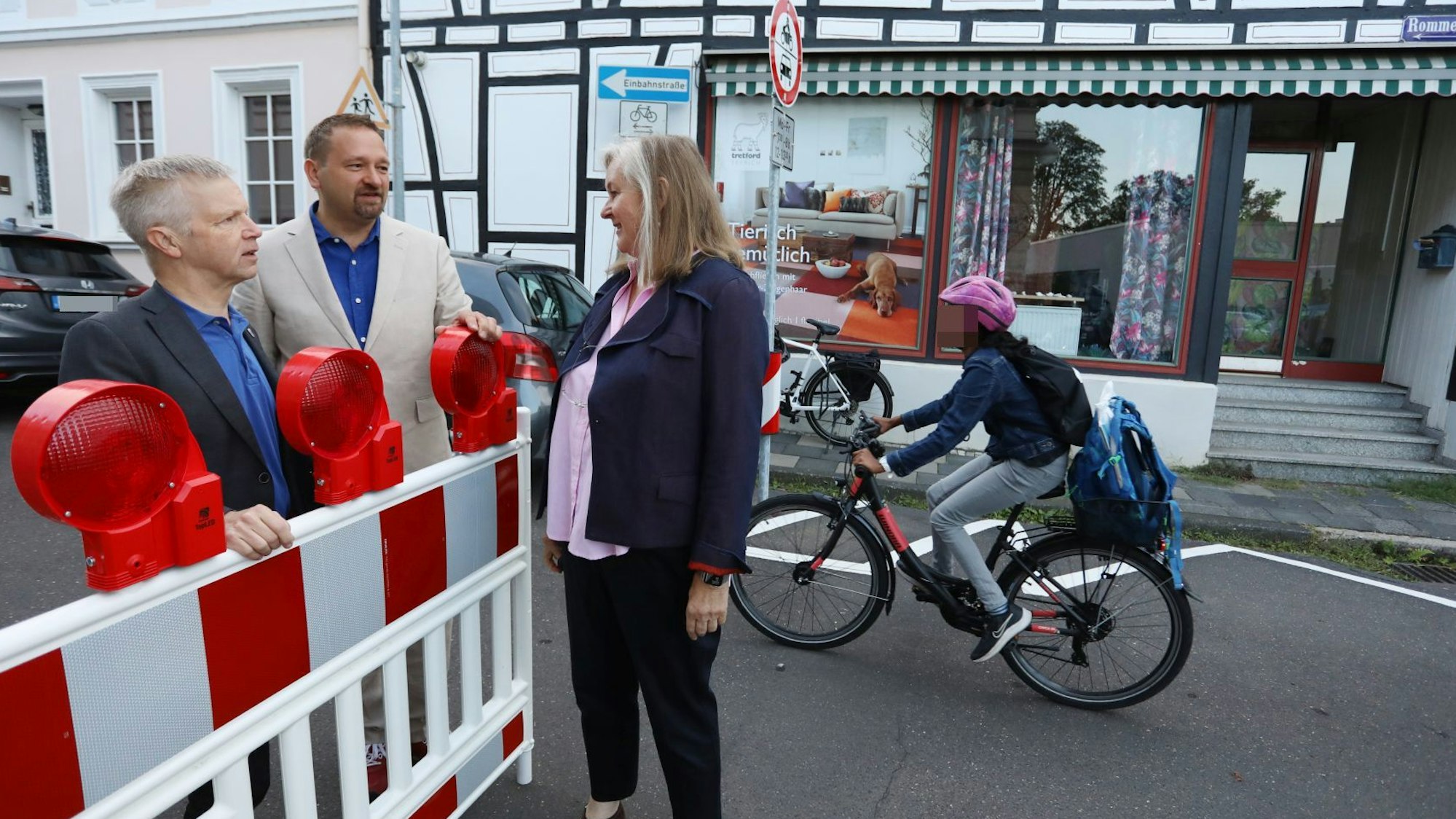 Zwei Männer und eine Frau stehen an einer rot-weißen Sperrbake, im Hintergrund fährt ein Schüler auf dem Fahrrad vorbei.