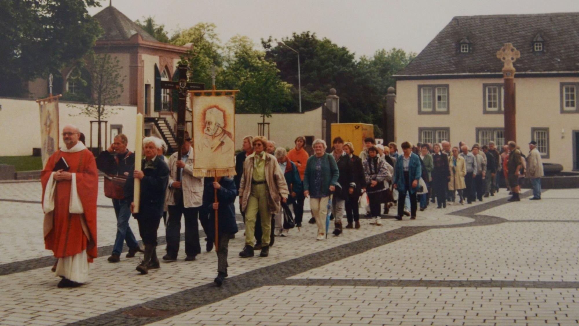 Hinter einem Priester zieht die Gruppe mit Kreuz, Kerze und Fahne über einen gepflasterten Platz.