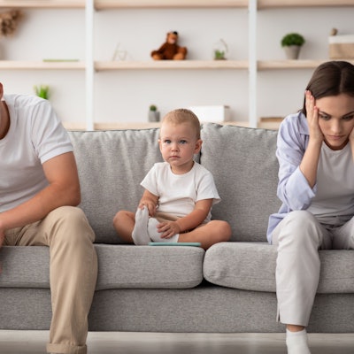 Ein junges Paar mit Kleinkind sitzt auf dem Sofa. Vater und Mutter stützen ihre Köpfe in die Hände.