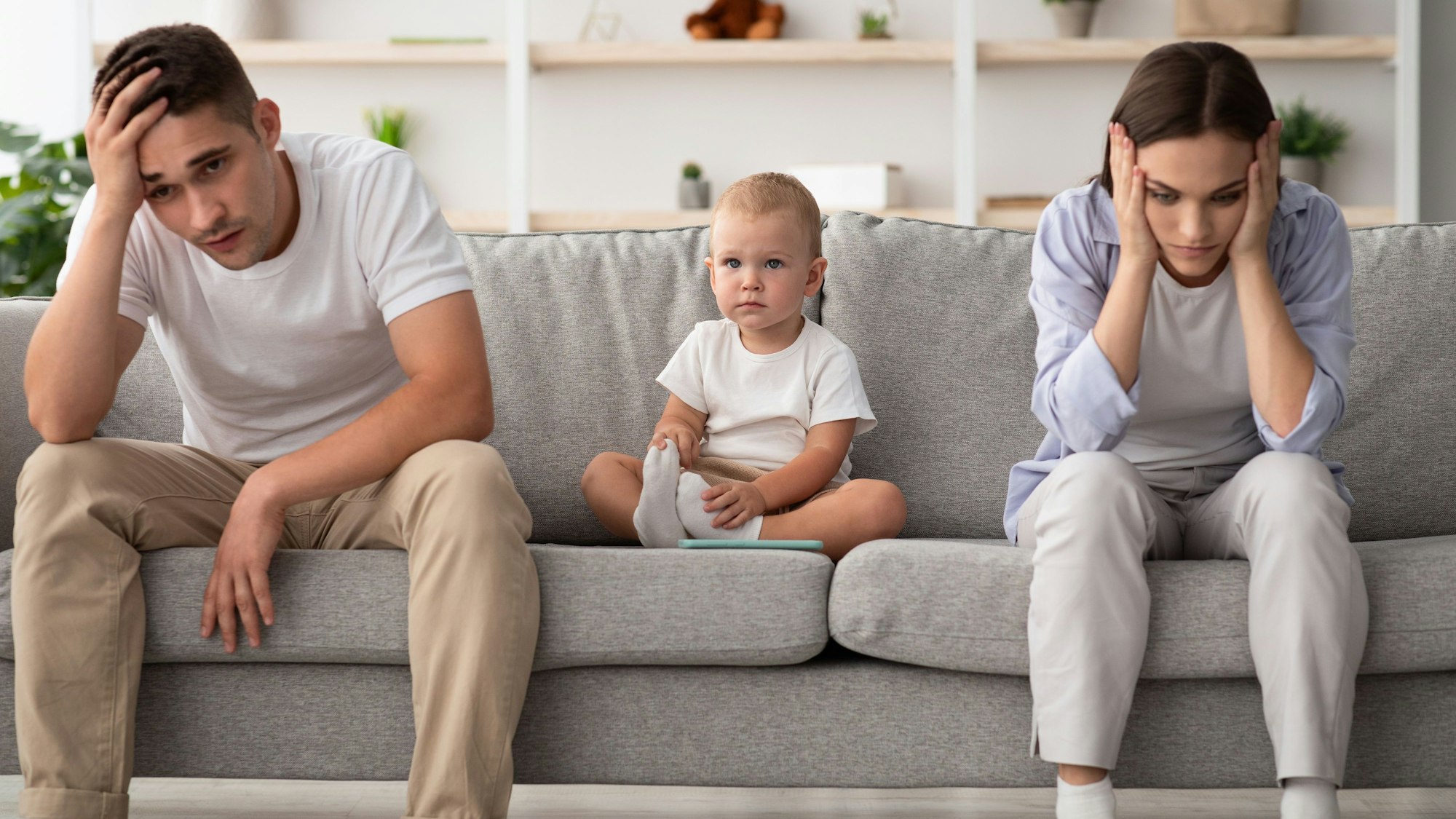 Ein junges Paar mit Kleinkind sitzt auf dem Sofa. Vater und Mutter stützen ihre Köpfe in die Hände.