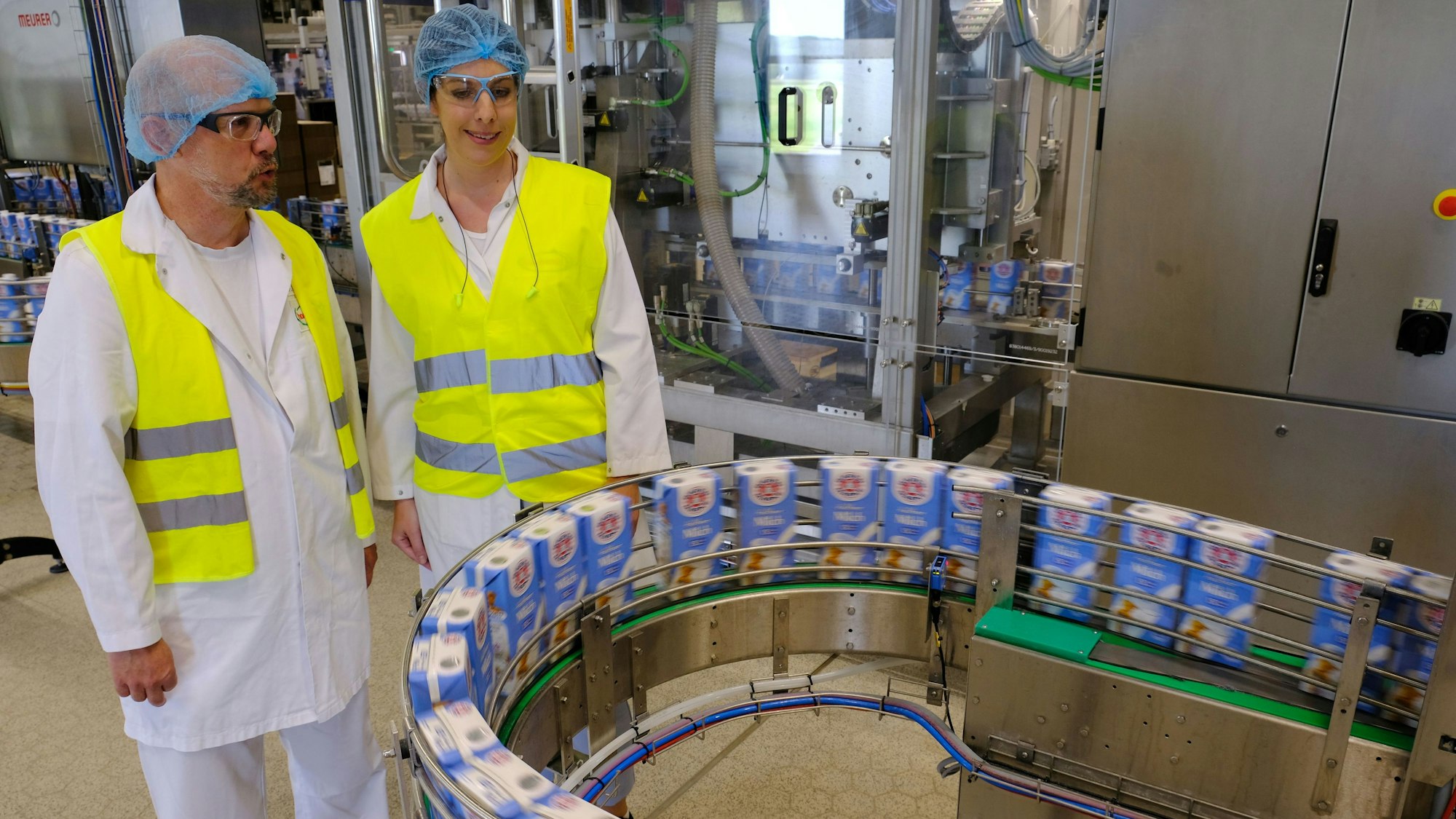 Zwei Mitarbeiter der Molkerei Hochwald in weißer Arbeitskleidung mit Haarnetz und gelber Sicherheitsweste stehen neben einem Förderband mit Milchverpackungen.