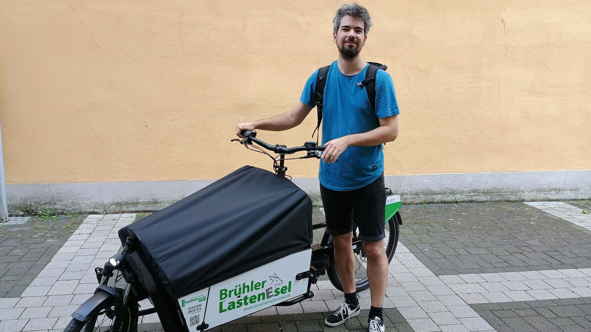 Auf dem Bild ist ein Journalist zu sehen, der mit einem Lastenrad der Kamera zugewandt steht. Er trägt ein blaues T-Shirt und eine schwarze kurze Hose. Am Lastenrad ist eine grüne Aufschrift auf weißem Grund "Brühler Lasten-Esel".
