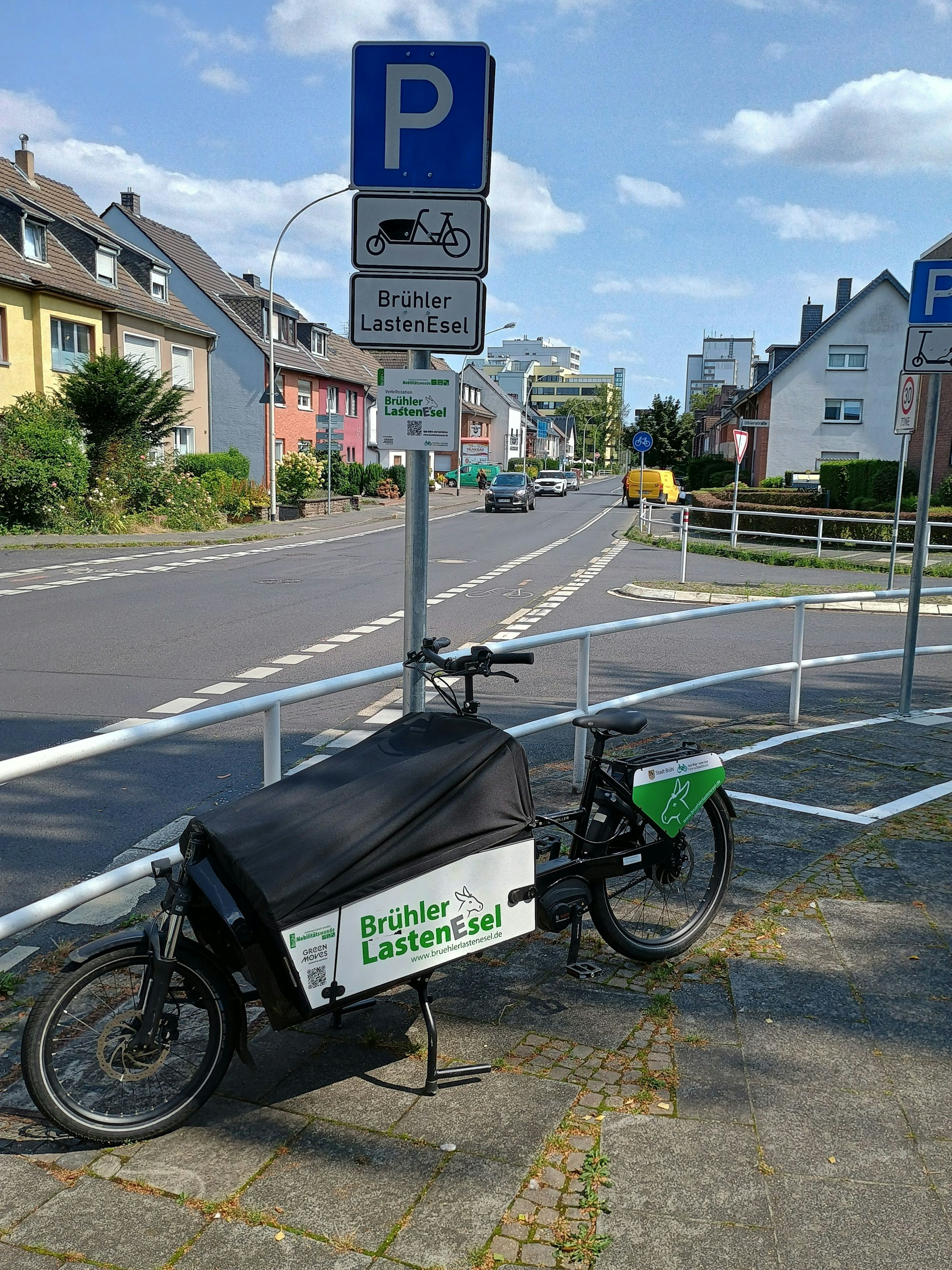 Auf dem Bild ist ein Lastenrad zu sehen. Ein Schild weist darauf hin, dass es gemietet werden kann. Im Hintergrund die Straße Rodderweg.