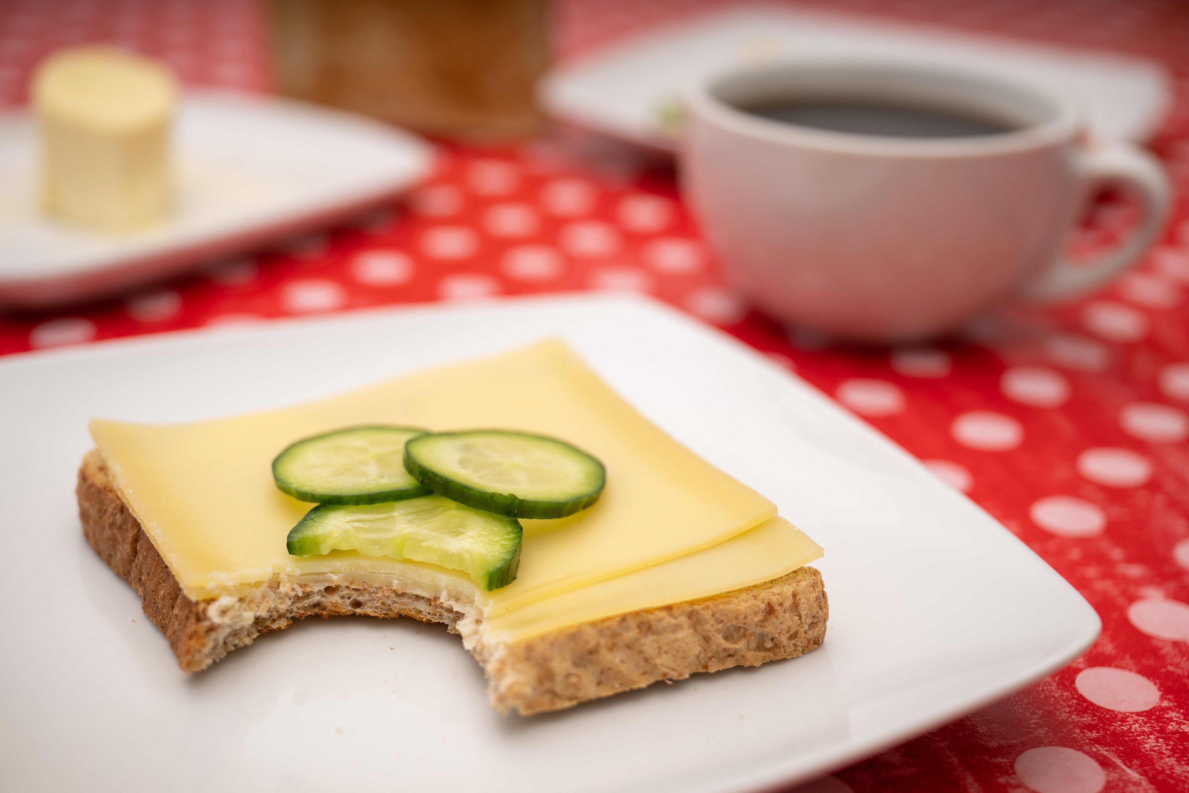 Ein Toastbroat mit Käse und Gurken liegt angebissen auf einem Teller. Dahinter steht eine Tasse Kaffee auf einer roten Tischdecke mit weißen Punkten.