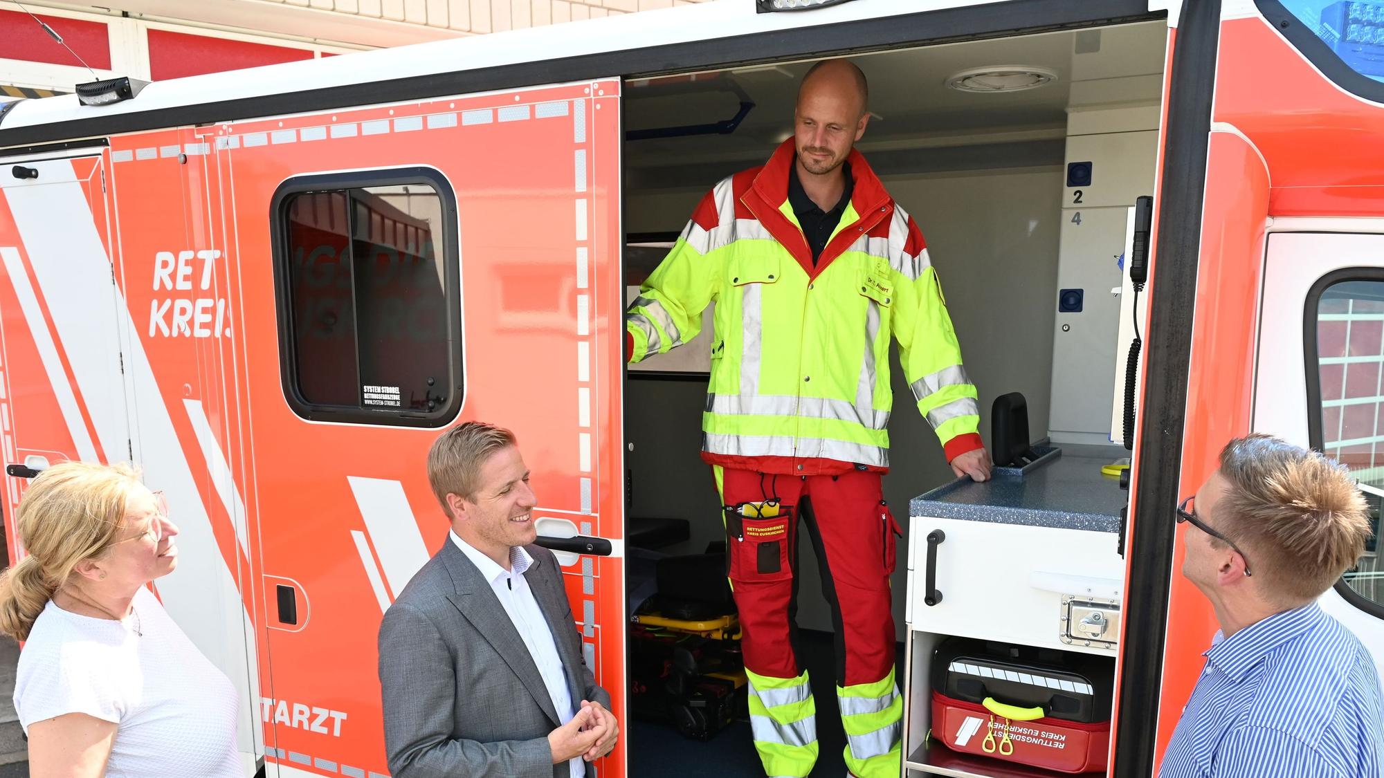 Tobias Ahnert trägt Rettungsdienst-Kleidung und steht in der offenen Tür eines Rettungswagens. Er unterhält sich mit Julia Baron, Markus Ramers und Martin Fehrmann, die vor dem Rettungswagen stehen.