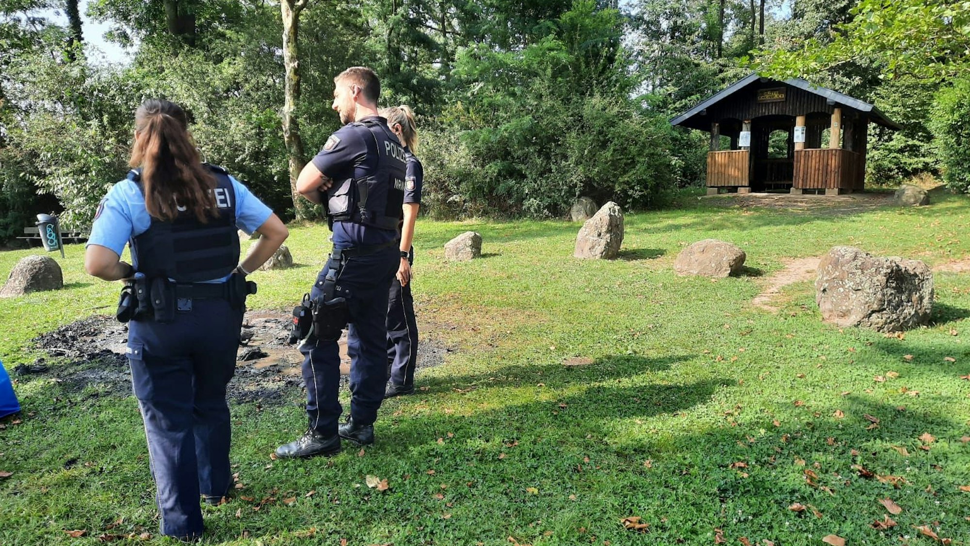 Drei Polizisten stehen neben einem verkohlten Stück Wiese, im Hintergrund ist eine Schutzhütte für Wanderer.