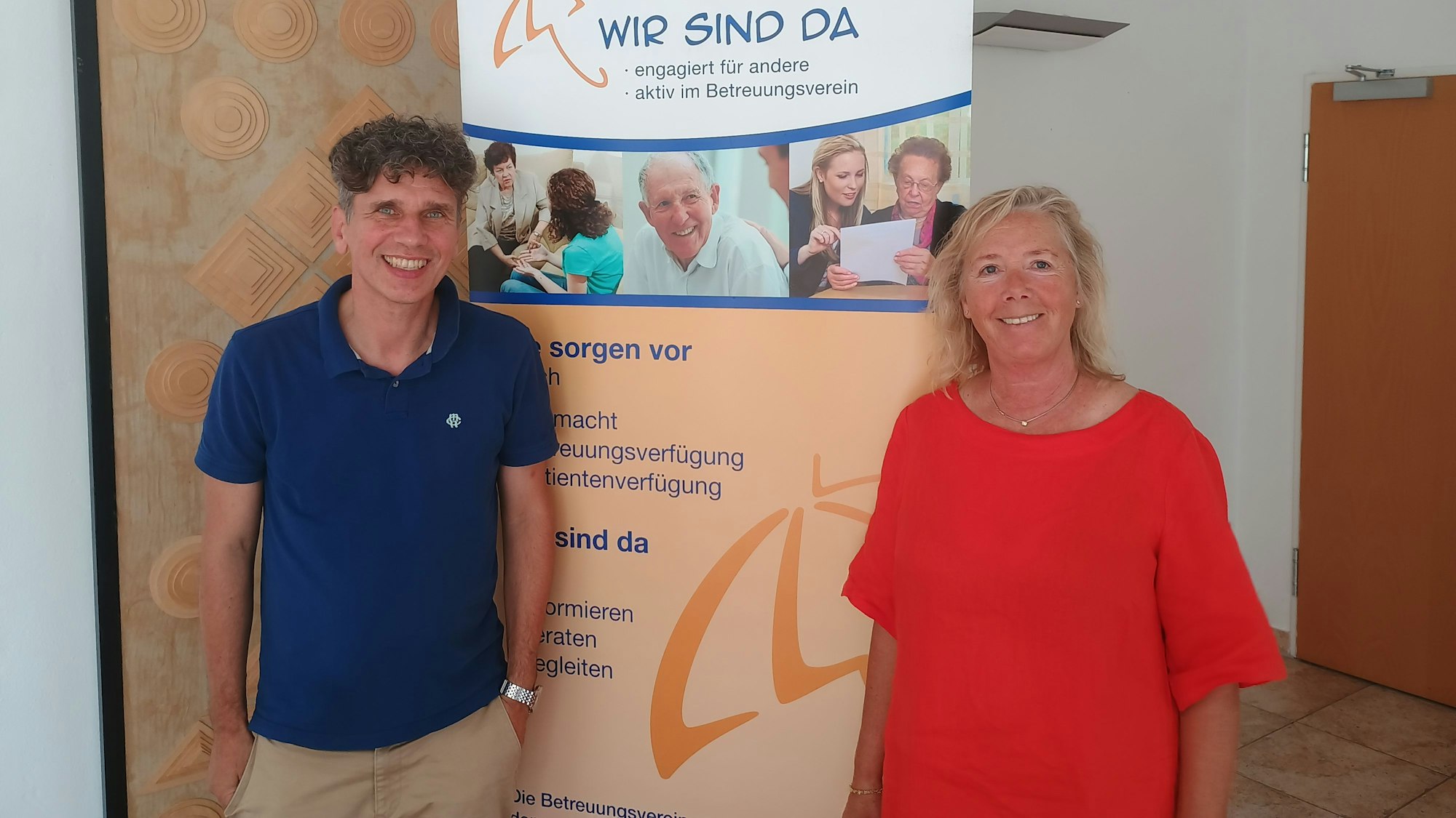 Frank Nixdorf steht links im Bild und trägt ein blaues Polo-Hemd. Rechts steht Anette Gövert, sie trägt ein rotes Kleid. Hinter den beiden ist ein Banner aufgestellt, auf dem die Aufschrift steht: „Wir sind da. Engagiert für andere, aktiv im Betreuungsverein.“