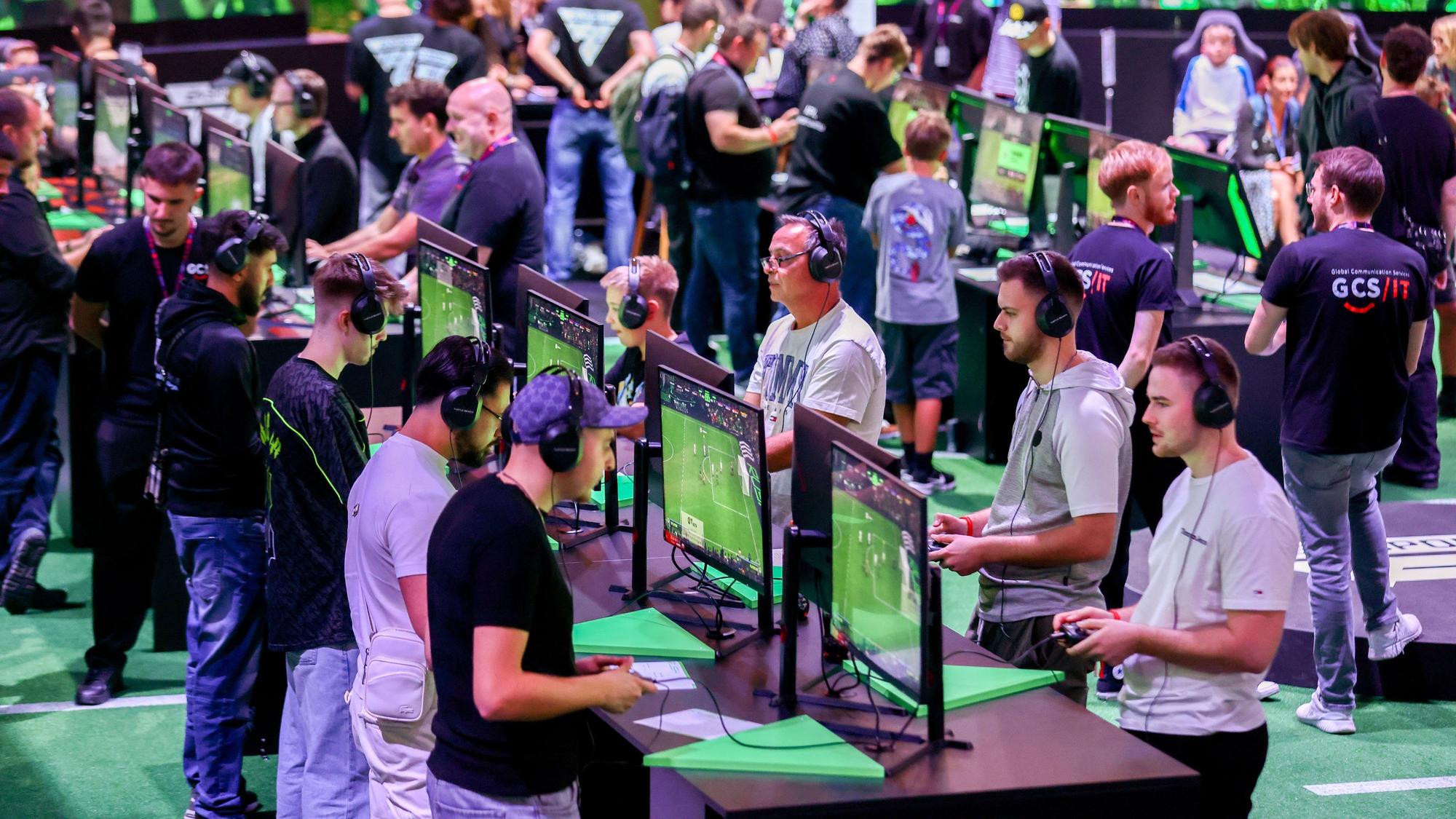 Bei der Gamescom 2024 stehen Gaming-Fans an Hochtischen, auf denen Computerbildschirme stehen. Sie haben Controller in der Hand und spielen Fifa.