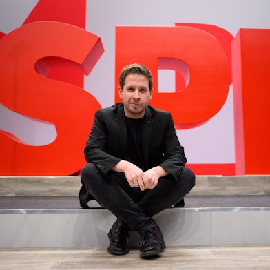 Kevin Kühnert, SPD-Generalsekretär, sitzt im Vorfeld des ordentlichen Bundesparteitags der SPD bei einem Hallenrundgang durch den Tagungsort vor einem SPD-Logo. (Archivbild)