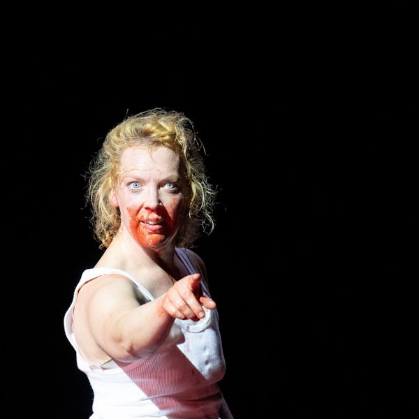 Lina Beckmann in "Laios" am Deutschen Schauspielhaus Hamburg. Die Schauspielerin trägt ein weißes Unterhemd und gestikuliert mit ausgestrecktem Zeigefinger, ihr Gesicht ist blutverschmiert.