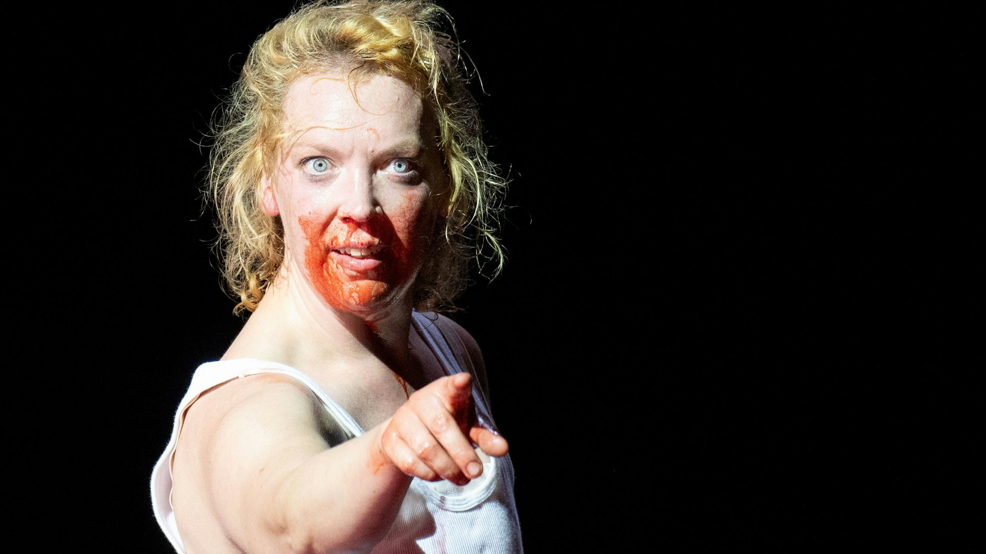 Lina Beckmann in "Laios" am Deutschen Schauspielhaus Hamburg. Die Schauspielerin trägt ein weißes Unterhemd und gestikuliert mit ausgestrecktem Zeigefinger, ihr Gesicht ist blutverschmiert.
