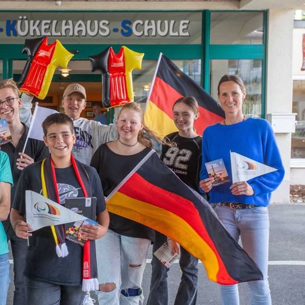 Mehrere Schüler und zwei Betreuerinnen stehen mit Deutschlandflaggen und weiteren Fan-Artikel in Deutschland-Farben vor einer Schule.