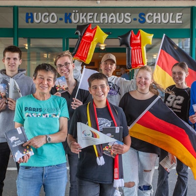 Mehrere Schüler und zwei Betreuerinnen stehen mit Deutschlandflaggen und weiteren Fan-Artikel in Deutschland-Farben vor einer Schule.