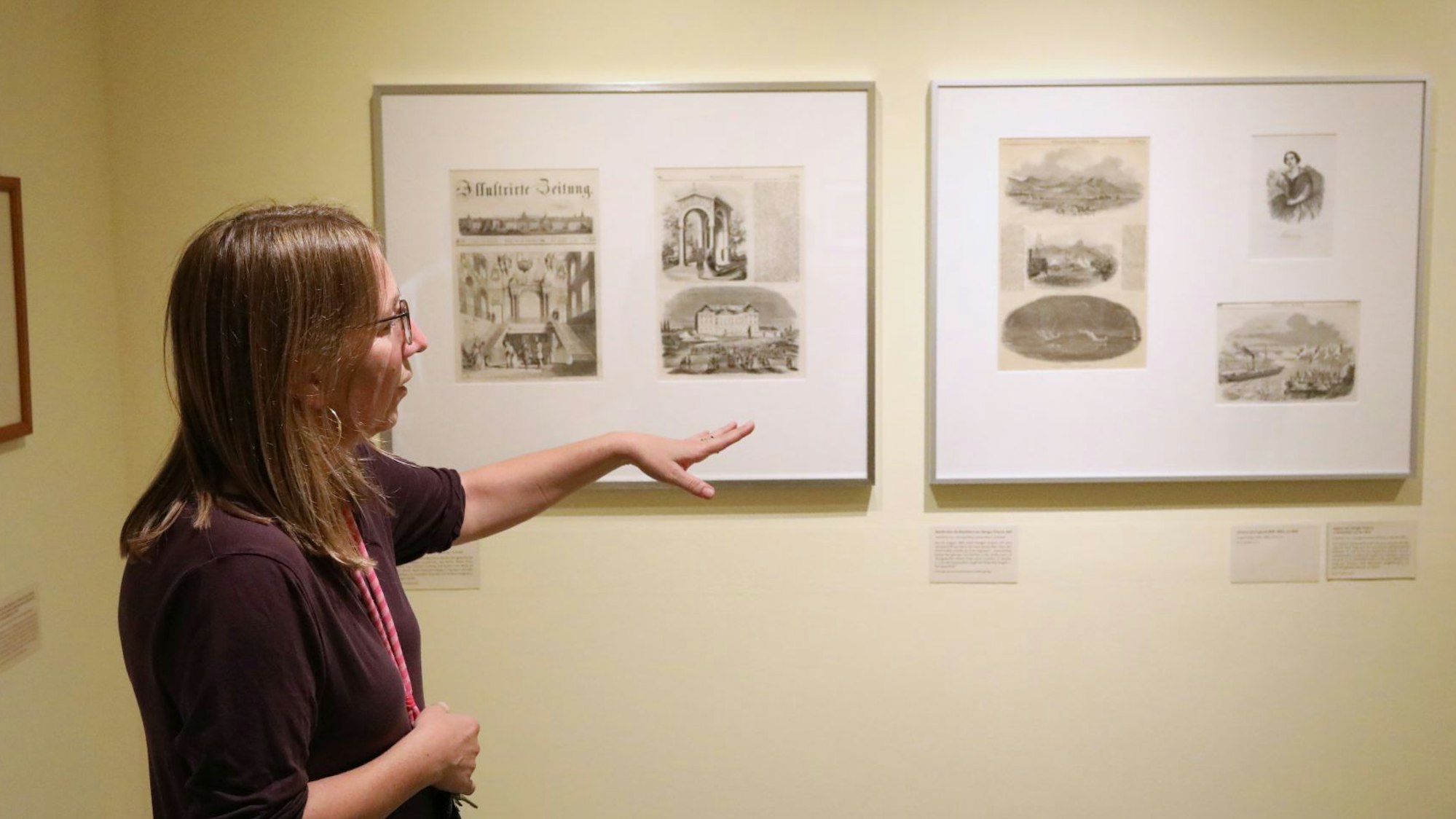 Eine Frau zeigt auf Bilderrahmen mit historischen Berichten.