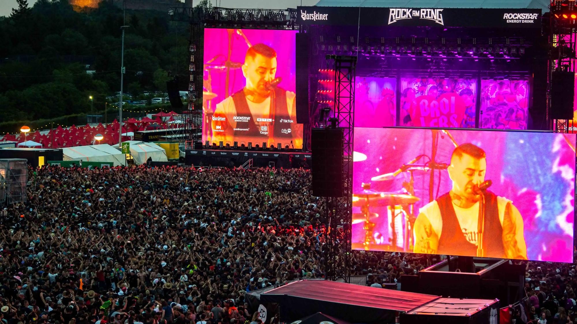 Menschenmassen haben sich in der Dämmerung vor einer Bühne bei Rock am Ring versammelt. Auf großen Bildschirmen sieht man einen Mann in pinkes Licht getaucht und Gitarre spielend.