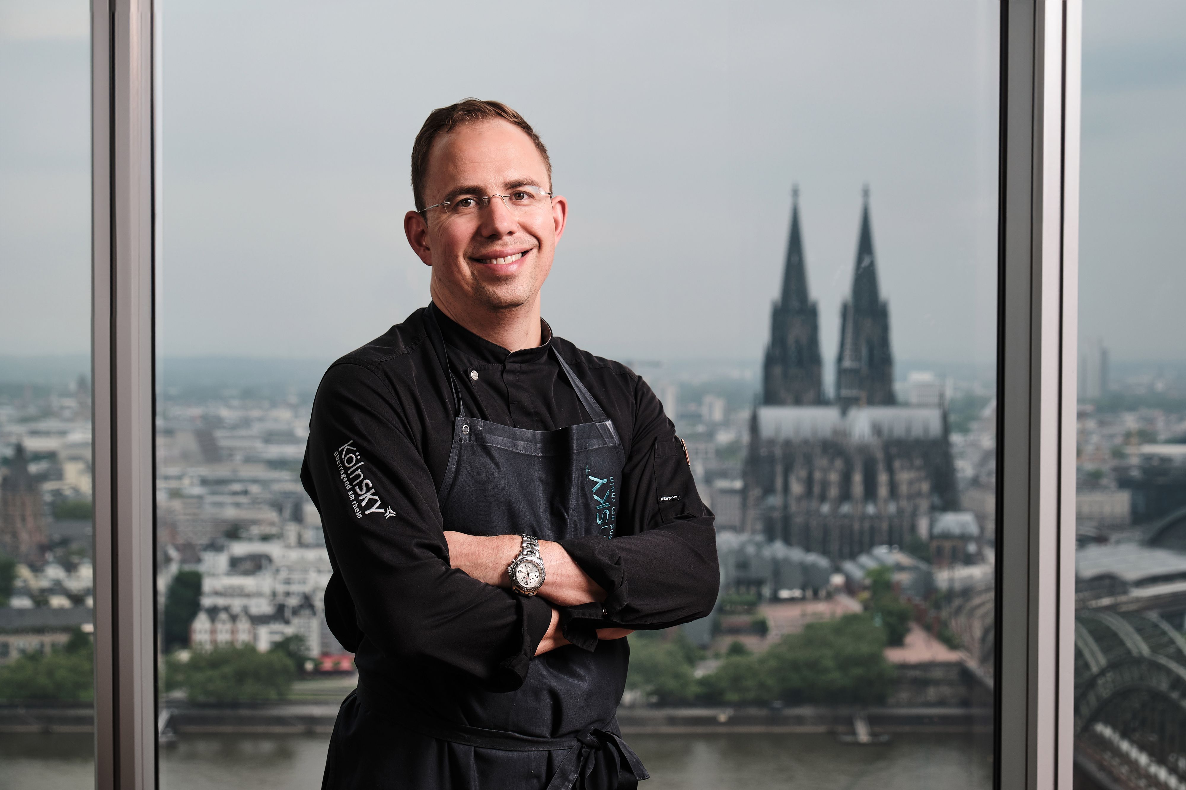 Ein Mann steht vor einem Fenster mit Schürze und dem Kölner Dom im Hintergrund.