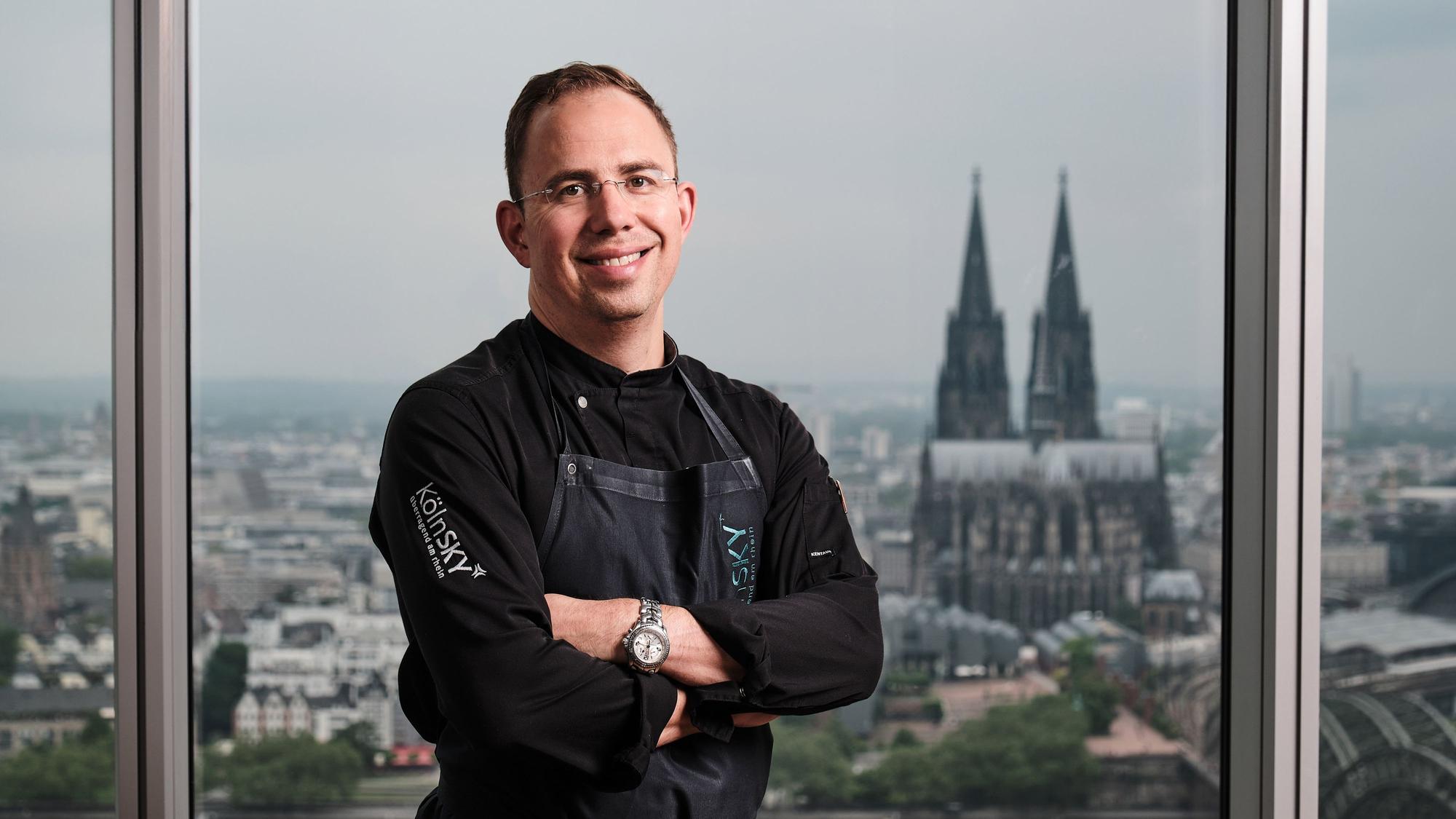 Ein Mann steht vor einem Fenster mit Schürze und dem Kölner Dom im Hintergrund.