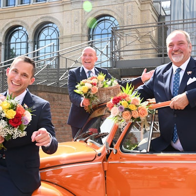 Das designierte Dreigestirn 2025 posiert im Anzug mit Blumenstrauß vor orangem Käfer.