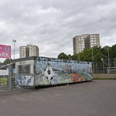 Im Vordergrund ist der abgesperrte Eingang zum Fußballplatz. Im Hintergrund sieht man die Hochhäuser.