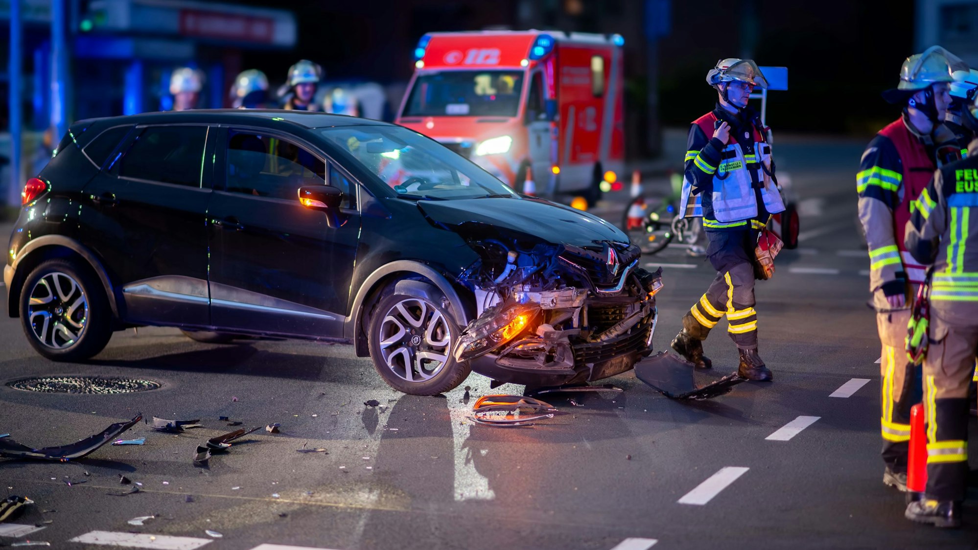 Das Bild zeigt ein am Unfall beteiligtes Auto. Im Hintergrund steht ein Rettungswagen, Feuerwehrleute begehen die Unfallstelle.
