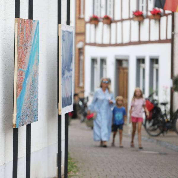Drei Gemälde nebeneinander, im Hintergrund eine Frau im blauen Kleid mit zwei Kindern auf der Straße.
