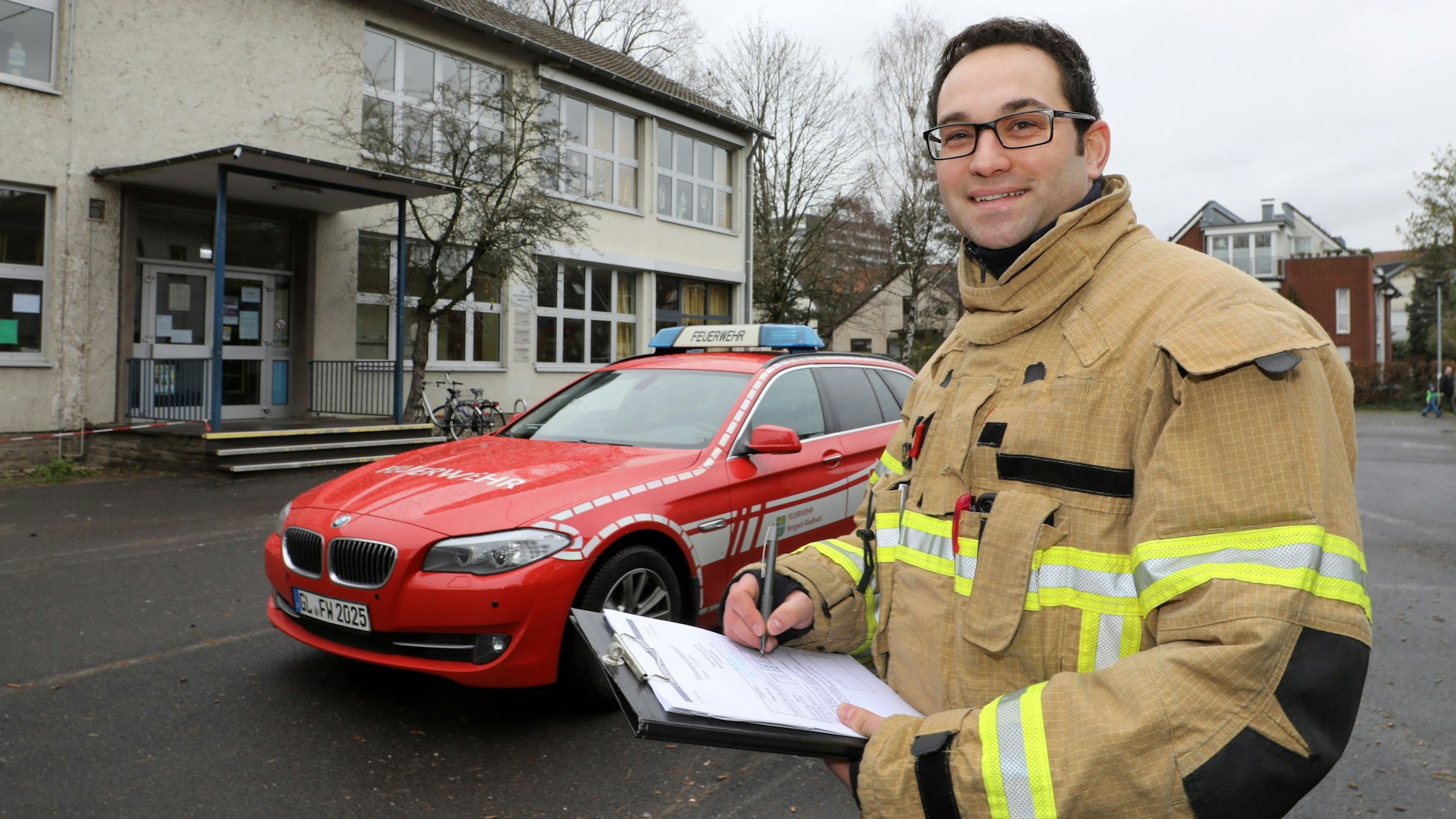 Löschzugführer Felix Vorndran steht mit einem Klemmbrett vor einem Feuerwehrauto auf einem Refrather Schulhof.
