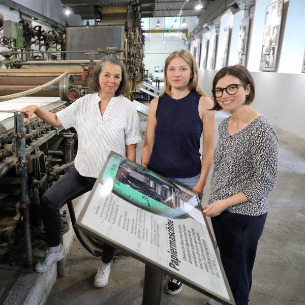 Die Museumsmacherinnen Sonja Nanko, Sophie Schmidt und Annette Schrick stehen an der Papiermaschine 4, die ab sofort wieder im Papiermuseum Alte Dombach zu sehen ist.