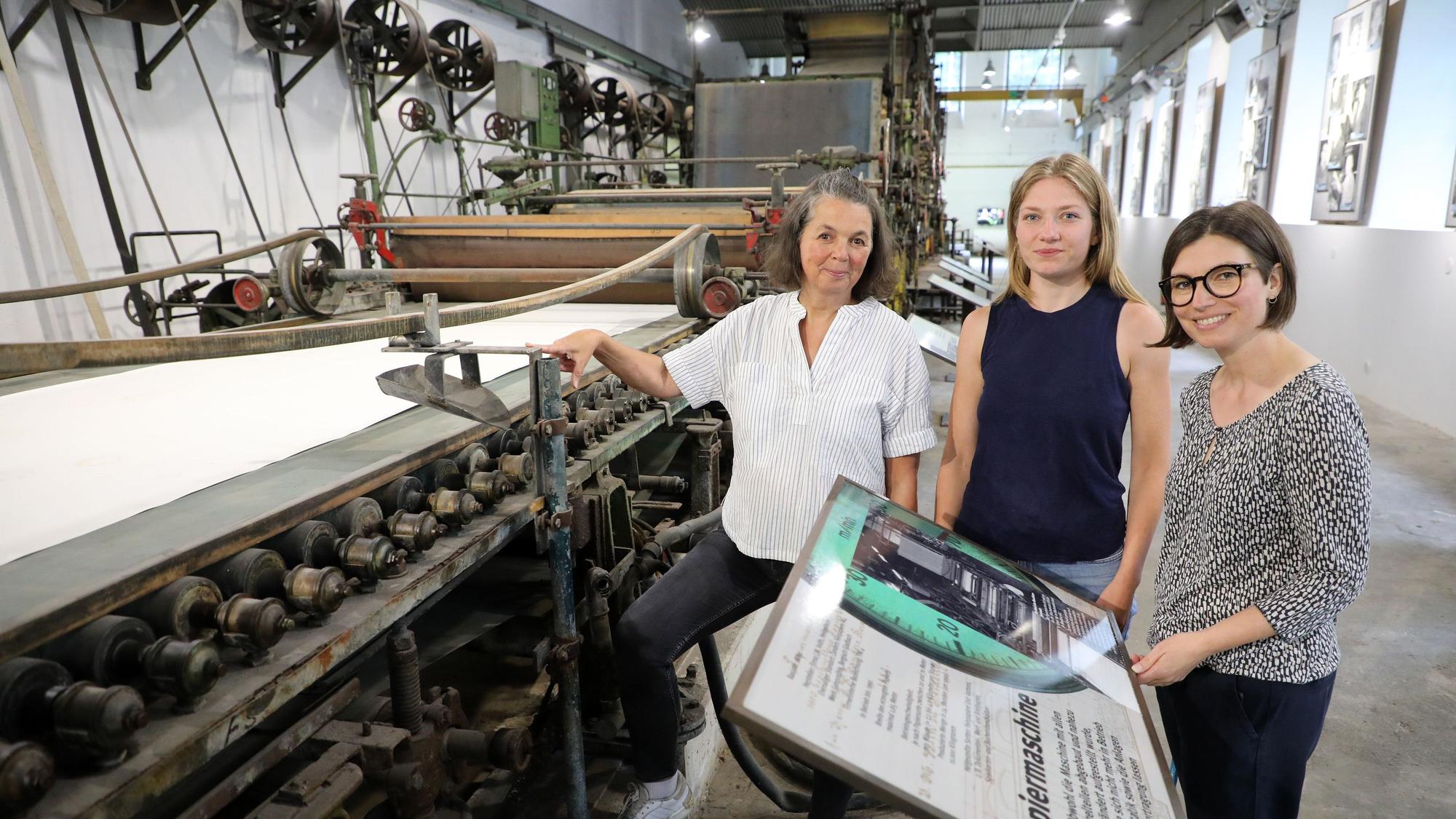Die Museumsmacherinnen Sonja Nanko, Sophie Schmidt und Annette Schrick stehen an der Papiermaschine 4, die ab sofort wieder im Papiermuseum Alte Dombach zu sehen ist.