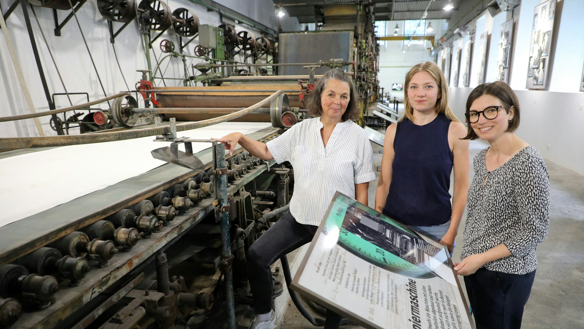Die Museumsmacherinnen Sonja Nanko, Sophie Schmidt und Annette Schrick stehen an der Papiermaschine 4, die ab sofort wieder im Papiermuseum Alte Dombach zu sehen ist.