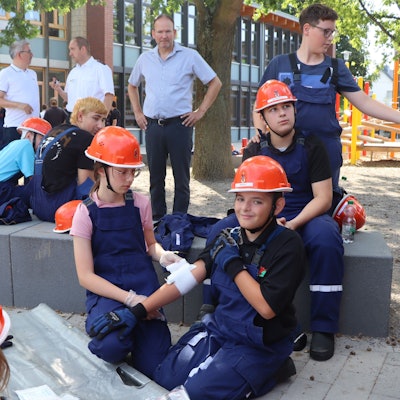 Auf dem Bild legt ein Mädchen eine Bandage um den Arm eines Jungen. Mehrere Kinder schauen zu. Sie alle tragen blaue Overalls und orangene Helme.
