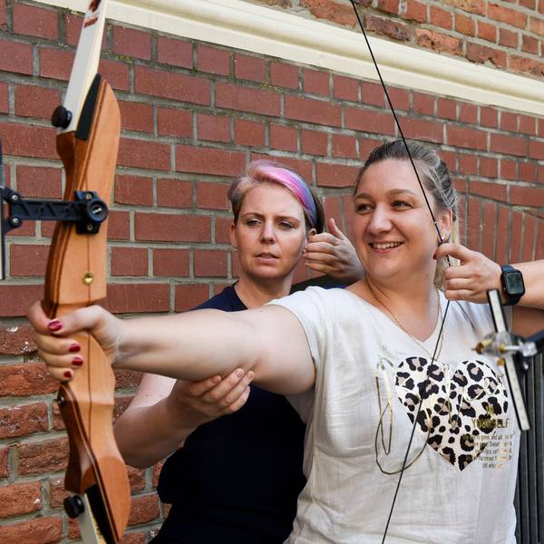 Auf dem Bild ist eine Frau zu sehen, die die Sehne ihres Bogens spannt. Eine Trainerin richtet den rechten Arm der Schützin.