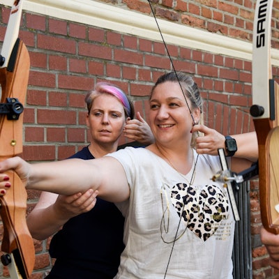 Auf dem Bild ist eine Frau zu sehen, die die Sehne ihres Bogens spannt. Eine Trainerin richtet den rechten Arm der Schützin.