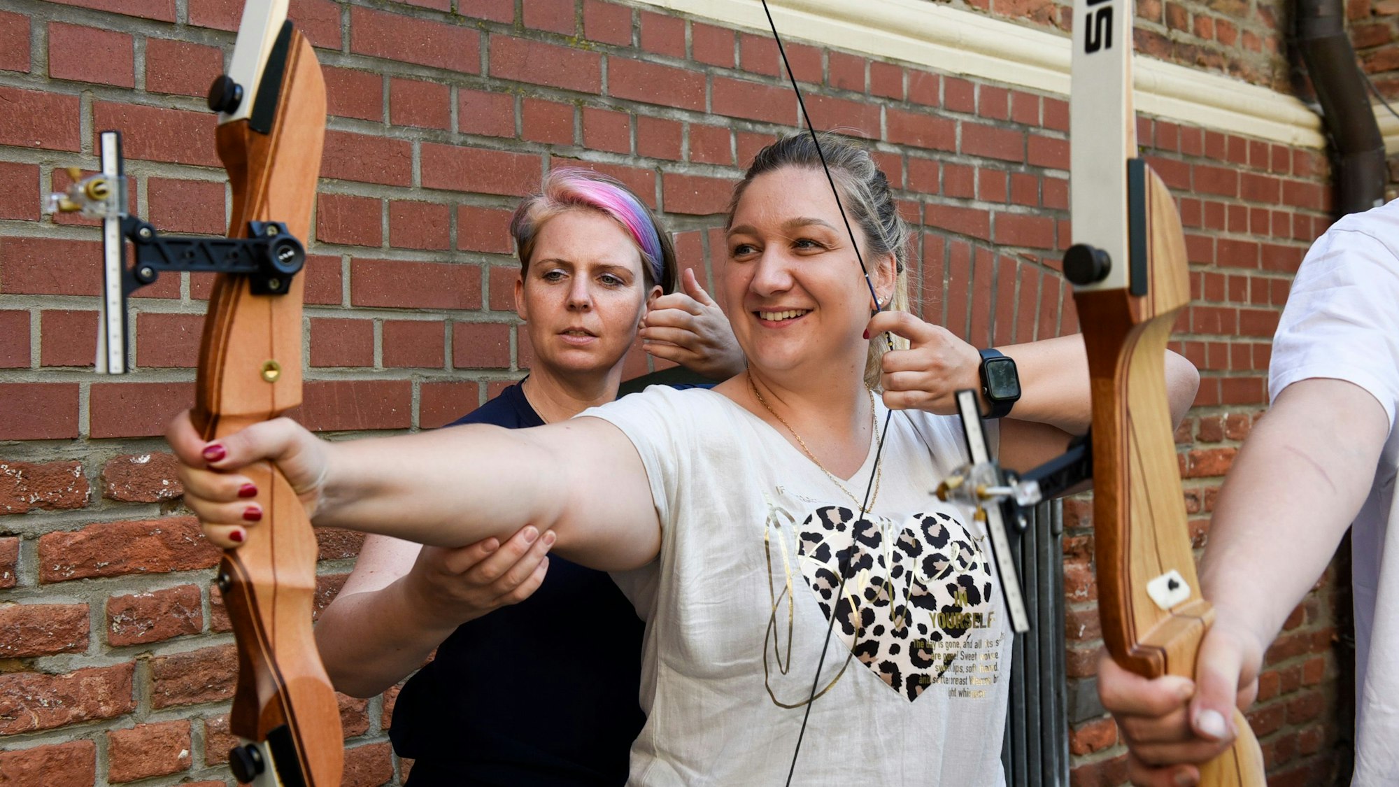 Auf dem Bild ist eine Frau zu sehen, die die Sehne ihres Bogens spannt. Eine Trainerin richtet den rechten Arm der Schützin.