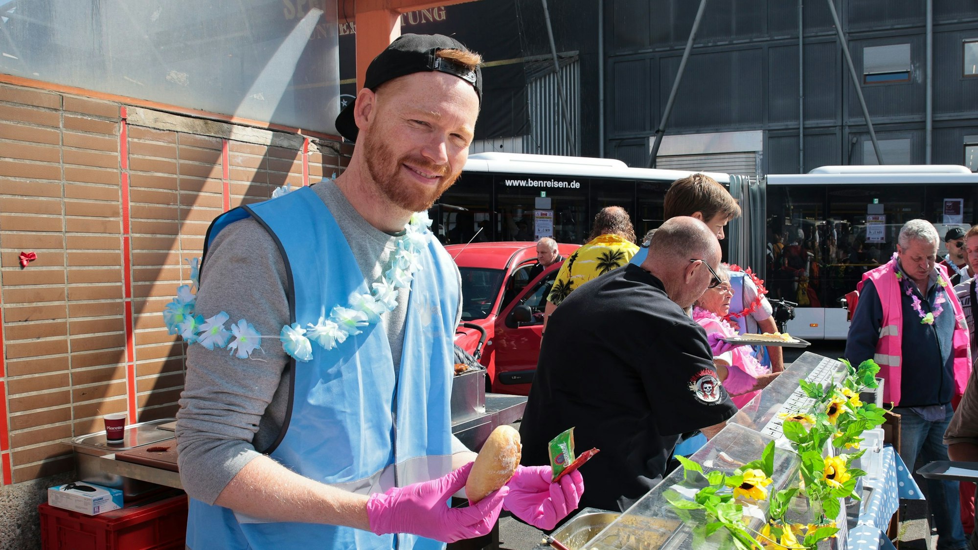 Nils Schreiber mit blauer Weste und Blumen-Kette an der Essensausgabe mit Brötchen und Saußenpäckchen in der Hand