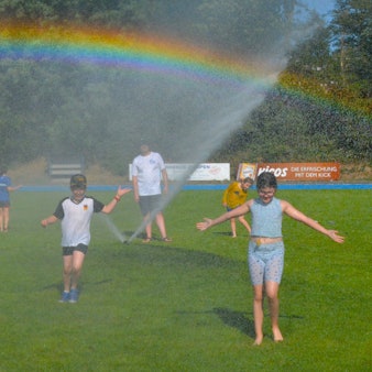 Kinder laufen über eine Wiese. Ein Rasensprenger sorgt für kühles Nass und einen Regenbogen.