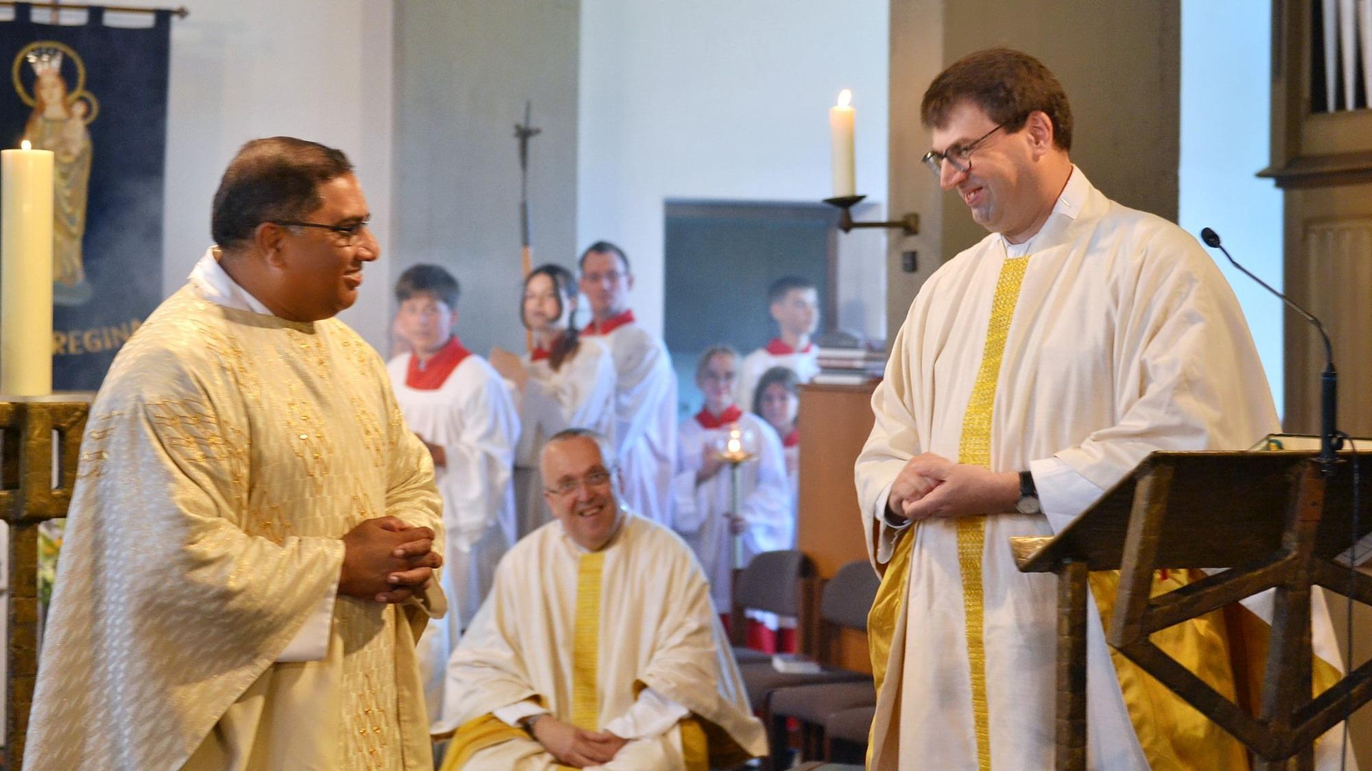 Zwei Männer in hellen langen Gewändern stehen sich in einer Kirche gegenüber und lächeln sich an, im Hintergrund sind Messdiener zu sehen.