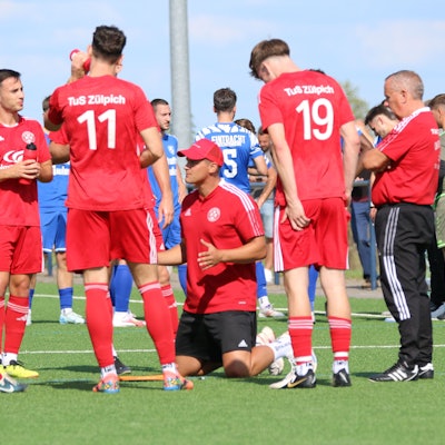 Zülpichs Trainer kniet während einer Trinkpause inmitten seiner Spieler auf dem Rasen.