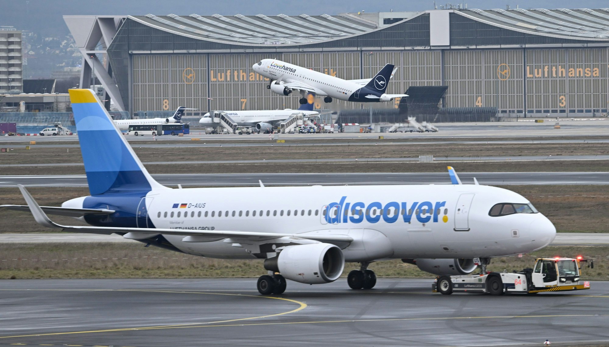 ARCHIV - 14.02.2024, Hessen, Frankfurt/Main: Während eine Maschine der Lufthansa startet, wird ein Passagierflugzeug der Discover Airlines am Frankfurter Flughafen von einem Schlepper gezogen. (zu dpa: «Gewerkschaften rufen Crews bei Lufthansa-Tochter zum Streik») Foto: Arne Dedert/dpa +++ dpa-Bildfunk +++