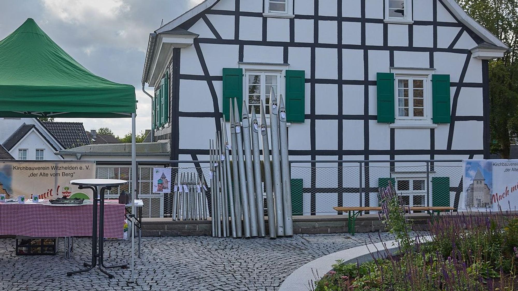 Orgelpfeifen stehen vor einem Fachwerkhaus.