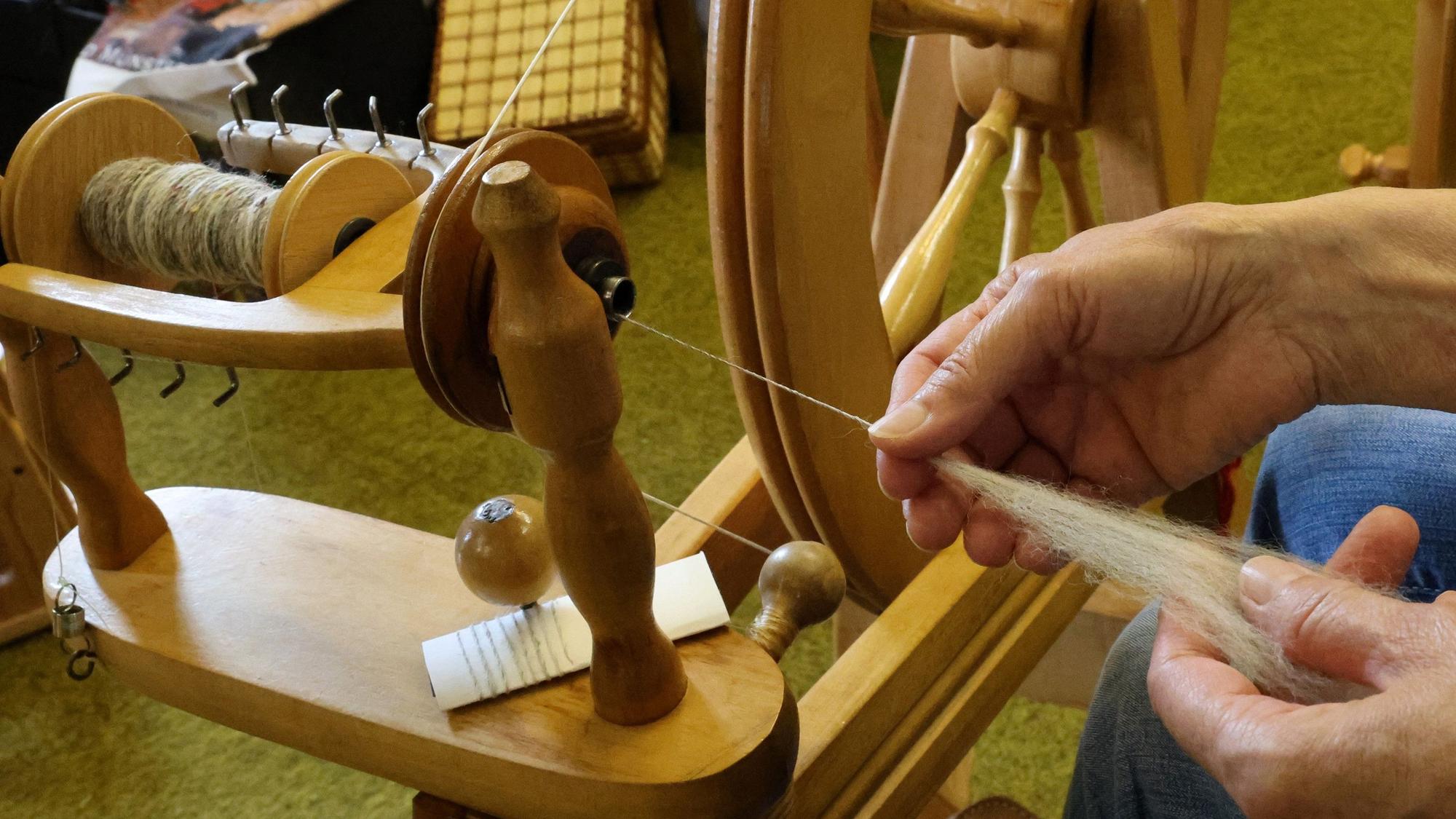 Eine Nahaufnahme zeigt die Arbeit mit Wollfasern, Spinnflügel und Spindel.