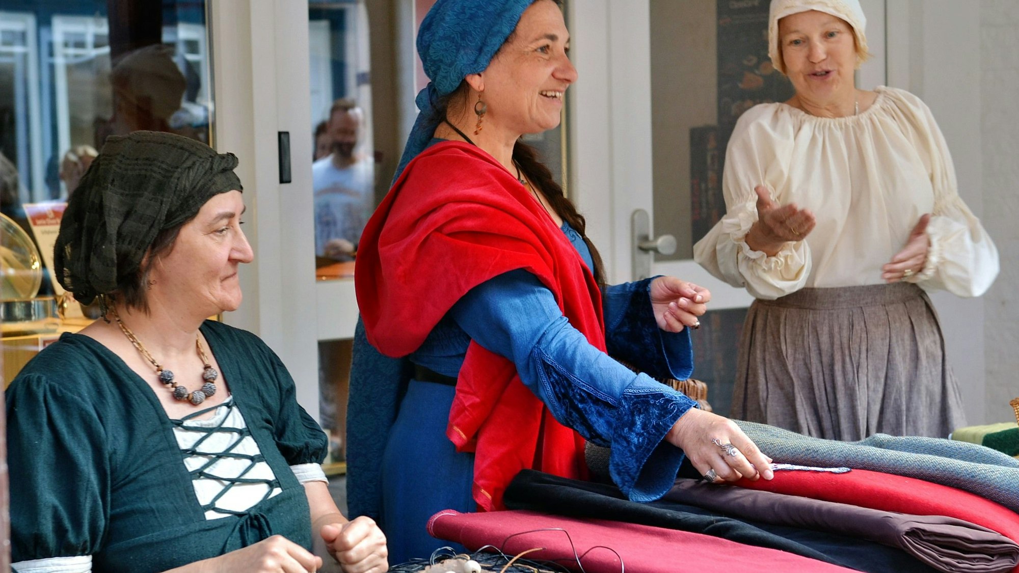 Drei in bunte Farben gewandeten Frauen mit Kopftüchern präsentieren Tuchwaren.