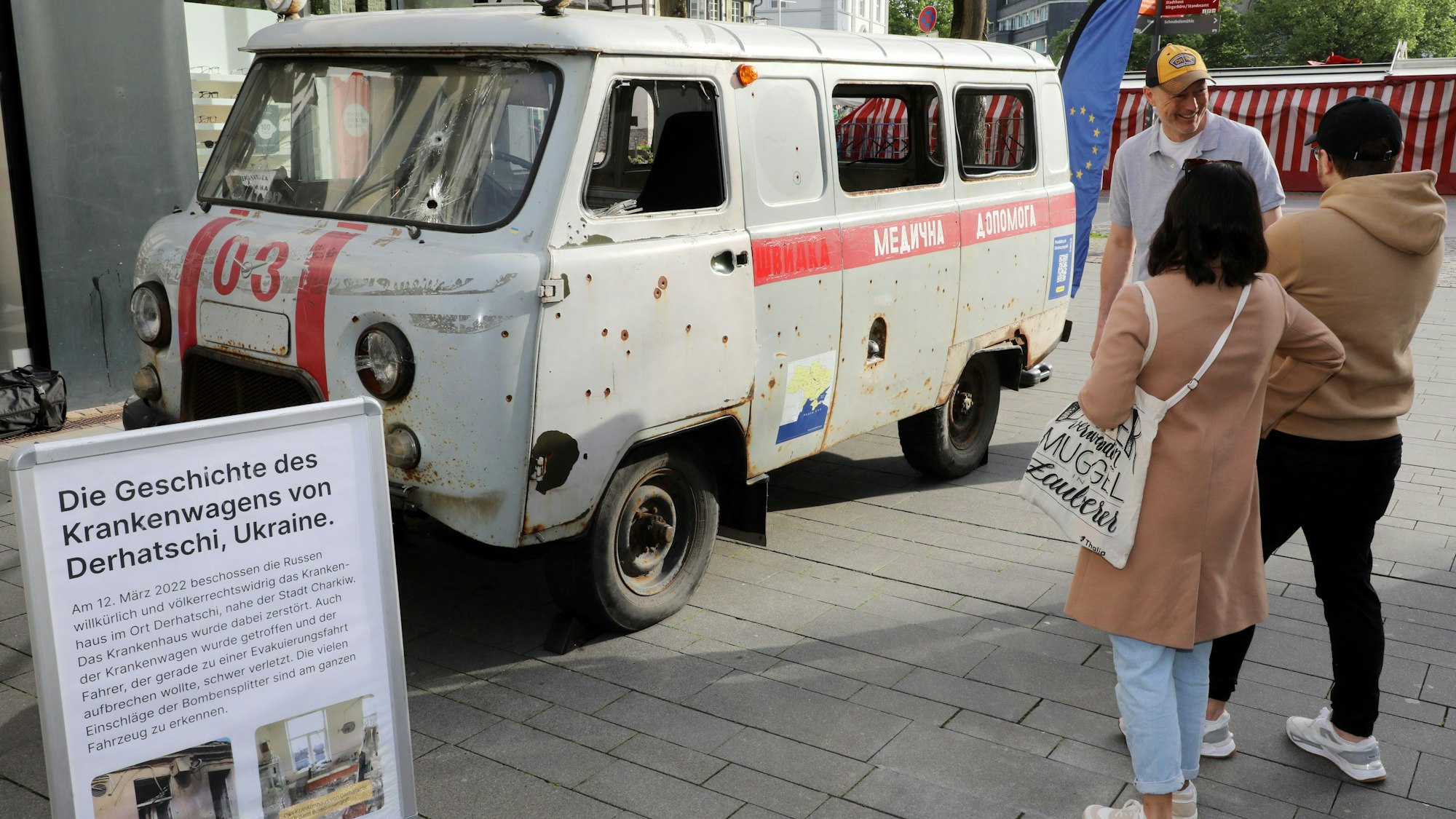 Menschen schauen sich einen beschossenen ukrainischen Krankenwagen in der Fußgängerzone von Bergisch Gladbach an.