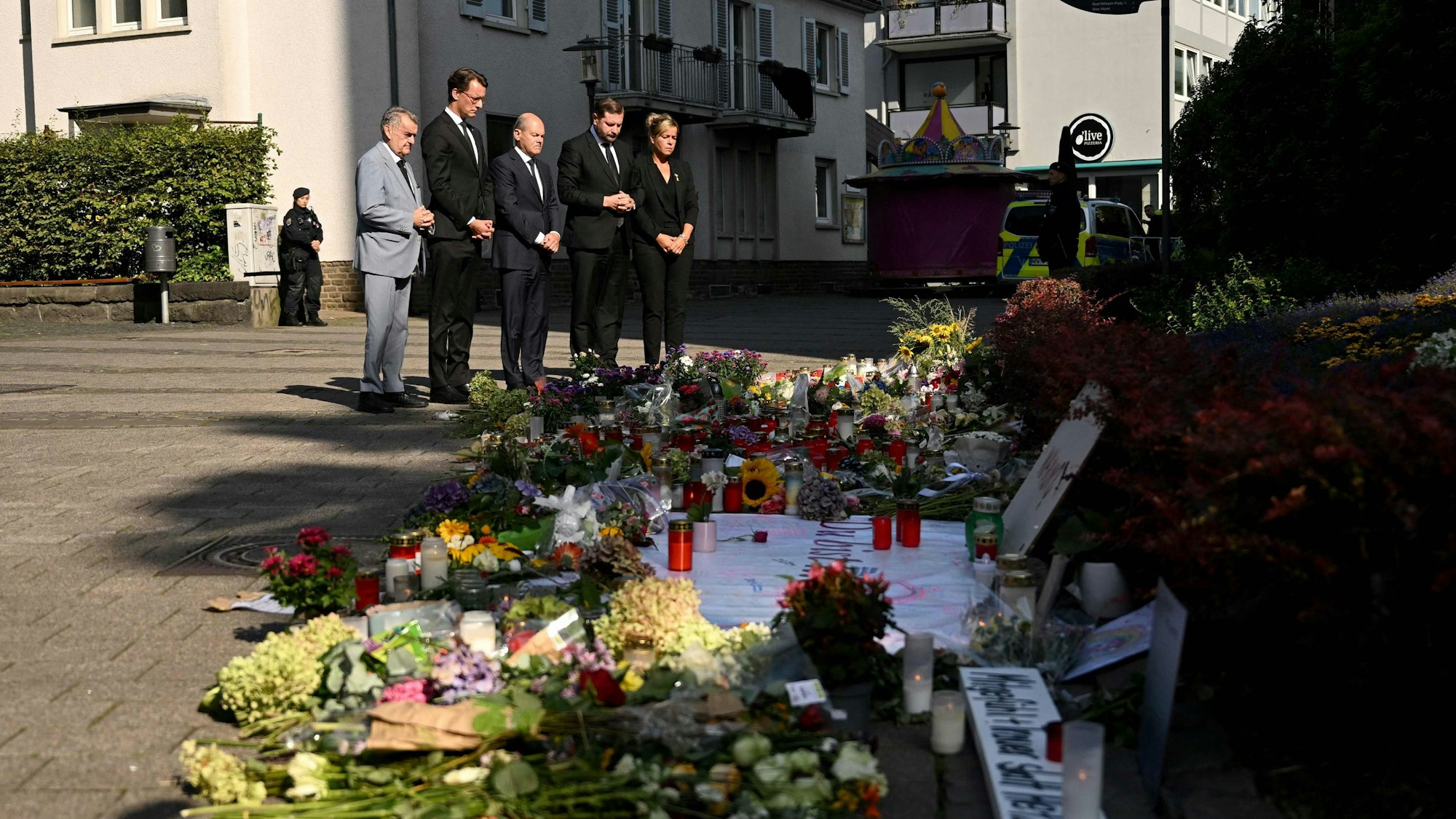 Mit dem Bundeskanzler ist auch die Spitze der Landesregierung nach Solingen gekommen. Neben Scholz (SPD) legen auch Ministerpräsident Hendrik Wüst, Innenminister Herbert Reul (beide CDU), Vize-Ministerpräsidentin Mona Neubaur (Grüne) und Oberbürgermeister Tim Kurzbach (SPD) stehen vor einem Blumenmeer am Tatort.